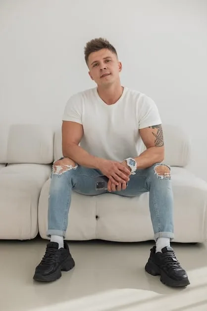 A man sits cross legged in a casual setting, wearing dark wash jeans, a white t-shirt, and stylish sneakers. He looks relaxed and confident.