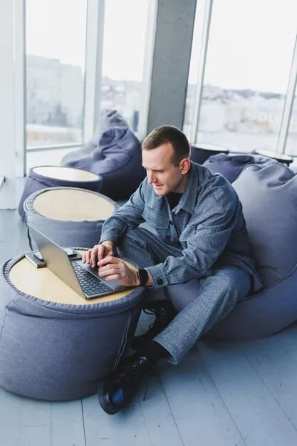 A man in a crisp, light-blue button-down shirt, looking relaxed and approachable, working on a laptop in a bright, modern office space.