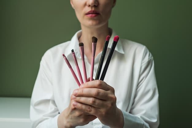 A focused shot of a person's hand holding a variety of eyeshadow brushes with different shapes and sizes. The background is a makeup palette with various eyeshadow colors. The brushes are clean and well-maintained, showcasing their quality and texture.