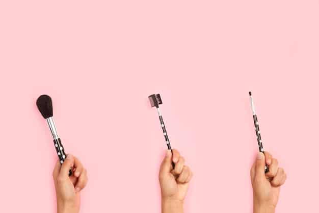 A hand holding a variety of makeup brushes, each with a different shape and size, against a blurred background of a makeup vanity. The focus is on the brush heads and their unique textures.