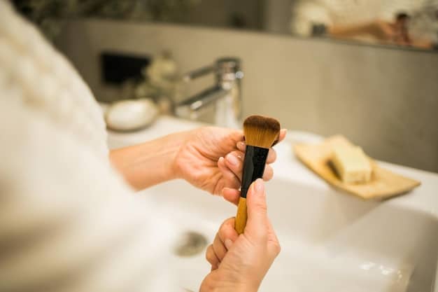 Close-up of a person gently swirling a foundation brush in soapy water, showing the makeup residue being released. The background is a clean bathroom sink.