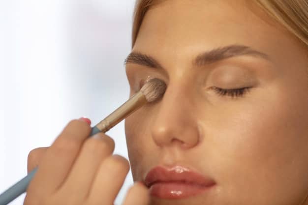 A detailed close-up of a woman applying monochromatic eyeshadow using a blending brush. The eyeshadow palette is visible, showing various shades of purple and lavender. The focus is on the blending technique and the seamless transition of colors on the eyelid.