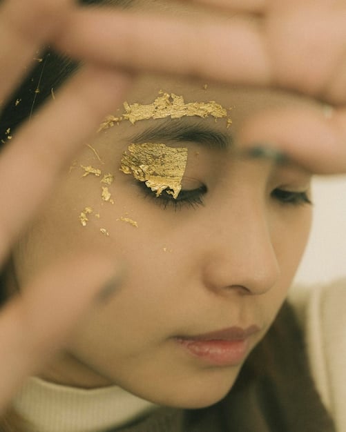 A detailed shot of a woman's eye with a gradient of gold and bronze eyeshadow, topped with chunky gold glitter applied to the center of the eyelid. The eyeliner is a sharp wing, and the lashes are long and voluminous.
