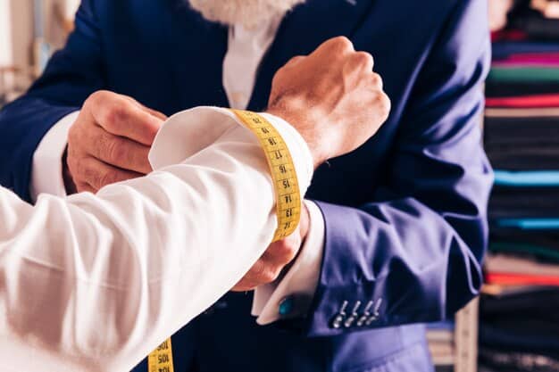 A close-up of a tailor's hands meticulously pinning the sleeve of a suit jacket on a mannequin, demonstrating precision and attention to detail. Tools like measuring tape and scissors are visible.