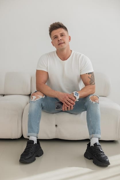 A man sits cross legged in a casual setting, wearing dark wash jeans, a white t-shirt, and stylish sneakers. He looks relaxed and confident.
